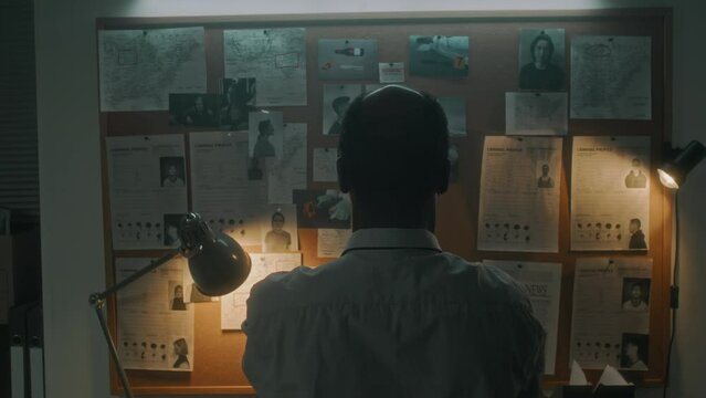 Medium rear shot of African American male detective standing in office, examining crime map with photos of suspects, victims body at murder scene, evidence, searching for motive