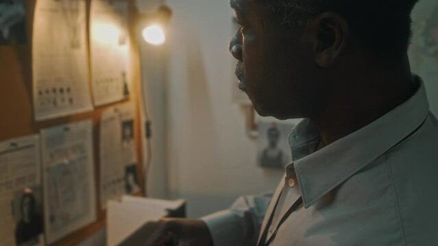 Medium close-up tilt shot of African American investigator working in office, pouring cup of coffee from glass jug and drinking, while staring at pin board with crime photos