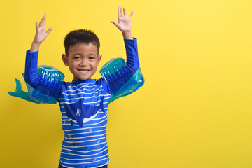 Portrait of a cute little Asian boy wearing a swimsuit posing with both hands raised isolated on a yellow background. Summer vacation concept. With Copy space