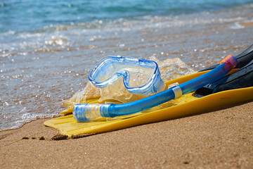 A pair of bright yellow flippers and a blue snorkel lie on the soft sand of a tropical beach. The turquoise ocean waves gently lap at the shore in the background.