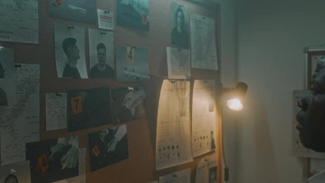 Close-up zoom out shot of African American detective pinning murder scene photos of victims body and evidence onto cork board on wall in office, with APBs for suspects, stepping back and examining it