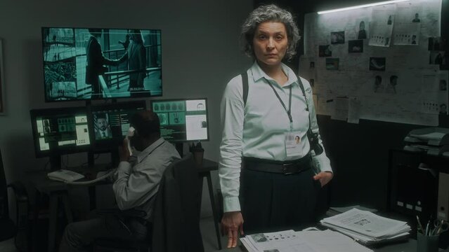 Medium full portrait of frowning mature Caucasian female intelligence agent, with ID badge, gun in holster, posing in office at table with evidence, black male colleague at CCTV desk talking on phone