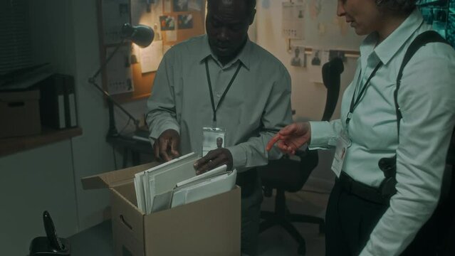 Medium full shot of Black male and Caucasian female police detectives bringing box of old case files from archive, laying out on table, searching for information while reopening unsolved investigation