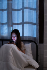 Asian sleepless woman in bed under blanket illuminated her cellphone using it for communication on social media and browsing internet at night with blue window light vertical style