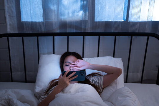 Asian young woman lying in bed at night holding a smartphone and covering her eye from the screen brightness of blue light unhealthy lifestyle before sleep