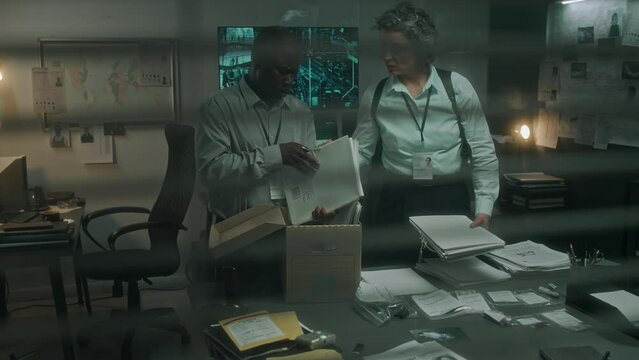 Medium full shot filmed through window blinds of African American male and Caucasian female law enforcement officers looking for information on current case in old archive files taken from storage
