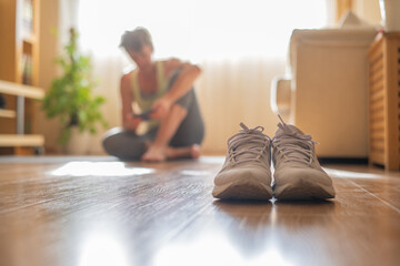 Home Workout Concept. Close-Up Of White Sneakers, In The Background Of A Mature Woman Sitting On A Yoga Mat Using A Mobile App For Her Morning Exercise Class