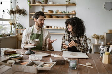 Mature man with beard in apron and curly-haired woman sculpting clay pottery together in a bright...