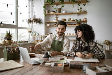 Bearded mature man and woman sculpting pottery in a studio while watching an online video course on a laptop