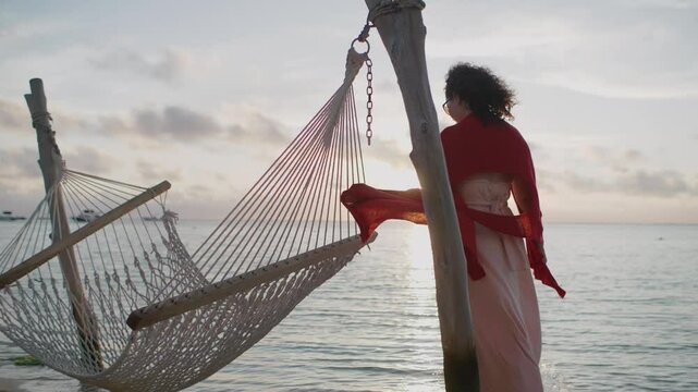 A woman draped in a shawl strolls around a hammock by the beach, basking in the serene sunset with boats visible in the background.