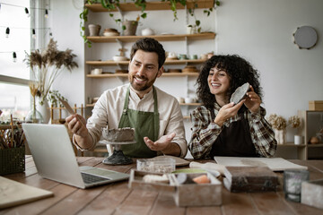 Bearded mature man and woman sculpting pottery, recording online video tutorial. Man creating vase, woman creating bowl or plate, both in aprons.