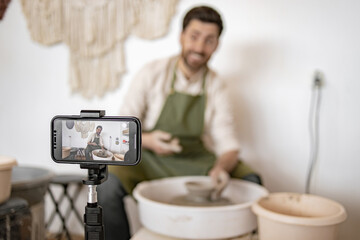 Young male potter sculpting pottery on a wheel while making a vlog. Capturing the artistic process on a smartphone camera.