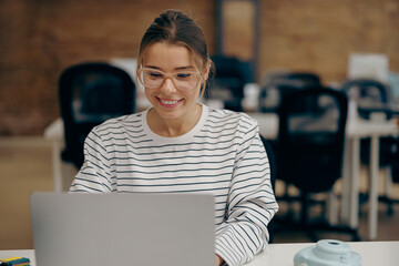 Young woman manager in eyeglasses working on laptop while sitting the desk in stylish office 