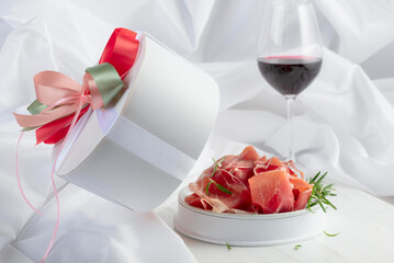 Prosciutto with rosemary in gift box and glass of red wine on a white wooden table.