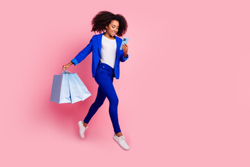 Full length photo of clever woman wear blue jacket fly with shopping bags look at smartphone empty space isolated on pink color background