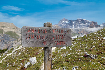 Wooden no camping prohibition sign marker on pole in italian dolomites in front of mountain range  against blue sky. Inscription in English, Italian and German language.