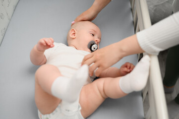 Caring mom has put baby with pacifier in cozy wooden crib, calms infant, pats head, top view. Nanny...