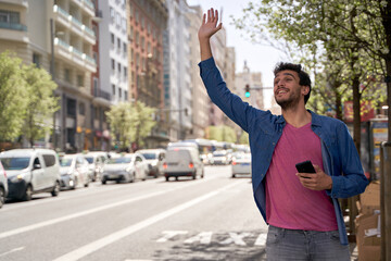 Young hispanic man stopping taxi with hand raised up using smartphone at street