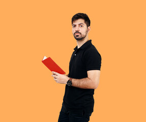 Confident Young Man Holding a Red Book Against an Orange Background. Boy with Book Wearing black shirt and black jeans. Young Male Student looking towards camera