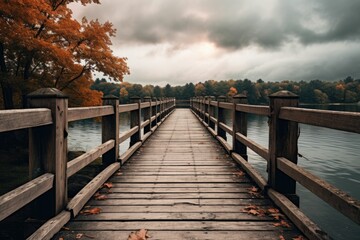 Fototapeta premium A wooden bridge stretches across a serene lake, flanked by vibrant autumn trees under a cloudy sky.