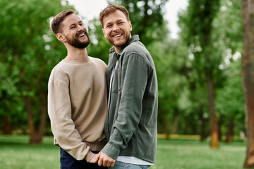 Obraz premium Two bearded men, an LGBTQ couple, laugh together in a green park on a sunny day.
