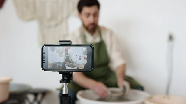 Adult male potter creating pottery on a potter's wheel while recording a video blog using a smartphone. Focus on ceramic art and social media content creation.