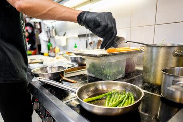 Close-up of a person’s hand in a black glove cooking green beans in a stainless steel pan on a stove, with a blurred kitchen background