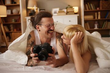 Tattooed man photographing his wife with a camera in bed