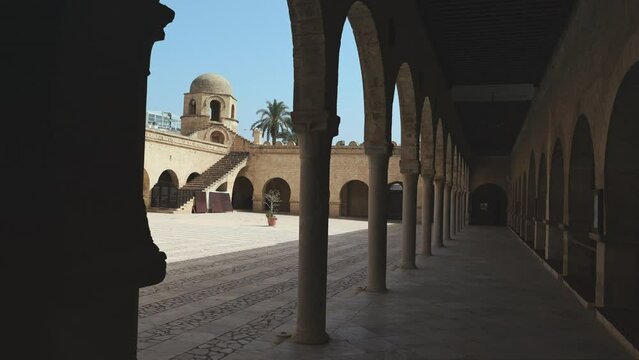 View from shaded archway gallery to sunlit courtyard of Great Mosque of Sousse and small domed corner tower rising against blue clear sky, showcasing traditional Islamic architecture. High quality 4k