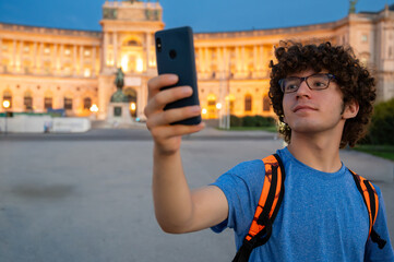 Vienna, Austria, August 17, 2022. A young Caucasian man with curly hair and glasses takes a selfie in front of the Hofburg Imperial Palace at sunset. The warm evening light enhances the imposing facad