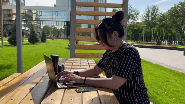 young brunette Asian woman student or freelancer working with laptop in the park outdoors on a sunny summer day