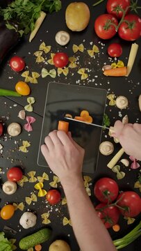 Overhead shot of crop Crazy cook cutting carrot on digital tablet, damaging the device and then stabbing knife at display on black table decorated, with vegetables and farfalle pasta Vertical shot.