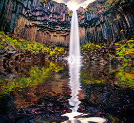 Huge basalt cliffs and Svartifoss (Black Fall) Waterfall reflected in the calm waters of small...