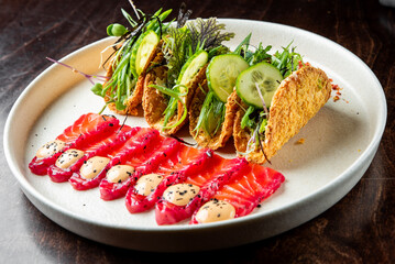 A gourmet plate featuring smoked salmon slices, garnished with herbs and sauce, alongside toast topped with greens and cucumber