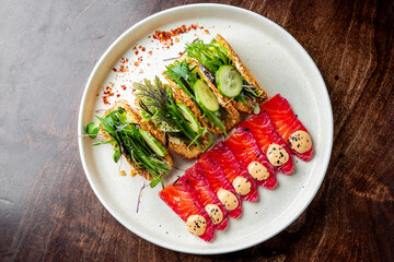 A gourmet plate featuring smoked salmon slices, garnished with herbs and sauce, alongside toast topped with greens and cucumber