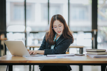 Young Asian professional business woman office worker analyst sitting at desk working on laptop thinking on project plan, analyzing marketing or financial data online, watching elearning webinar.