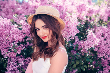 Young beautiful smiling woman in trendy summer clothes. Sexy carefree model posing in the street. Positive model near blooming flower lilac bush beaming. Summertime, pink colours. Red lips