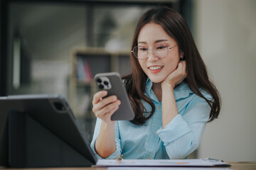 Fototapeta premium Beautiful Asian woman smiling happily working in modern office with documents, tablets and mobile phones on the table