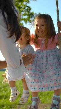 Happy brunette woman in a white dress swings her daughters on a rope swing in the middle of a green field. Two babes are laughing and smiling. Tree branch and blue sky at the bg. Slow motion. Vertical