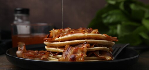 Pouring maple syrup onto delicious pancakes with fried bacon on wooden table, closeup