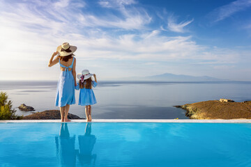 A beautiful mother and her daughter are standing by the pool and enjoying the summer sunset over the mediterranean sea