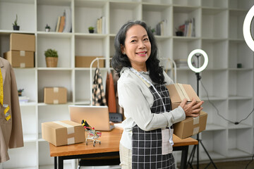 Confident senior woman small business owner holding parcel boxes of product and smiling to camera