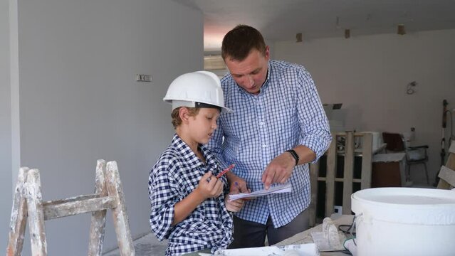 Father and his young son wearing protective hardhats discussing work plan in construction site in apartment.
