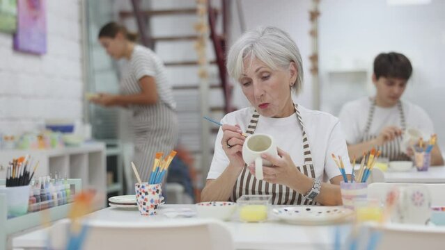 Interested elderly woman attending pottery class, enthusiastically painting on ceramic cup, creating unique custom design. Creative hobby concept