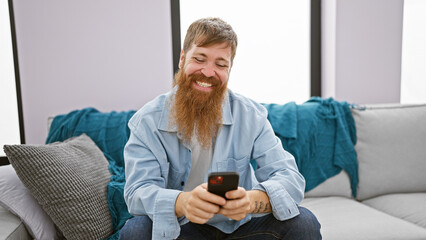 Handsome young redhead man cheerfully using his smartphone while sitting on the sofa in the living room of his home, spreading joy and positive vibes through texting and the online world