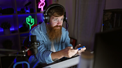 Intensely concentrated young redhead man steaming video game on smartphone in dark gaming room at night