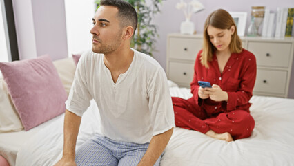 Obraz premium A contemplative man and a woman using a smartphone sit separately on a bed in a cozy bedroom, conveying a tension in their relationship.