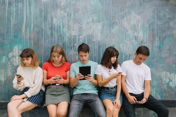 Group of teenagers sitting at the colored wall and using their mobile phones and gadgets
