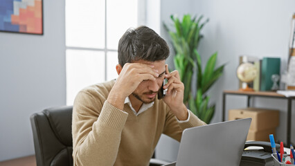 Stressed young hispanic man with a beard in an office using a laptop and phone, visually conveying professional tension.