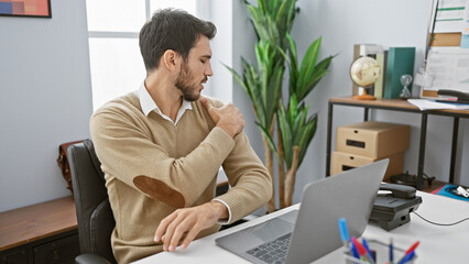 Hispanic man with beard in pain holding shoulder while sitting at office desk with laptop and indoor plant.